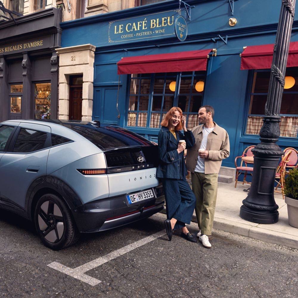 Blue Hyundai IONIQ 3&nbsp;parked on the street in front of a cafe with two people holding coffee cups.