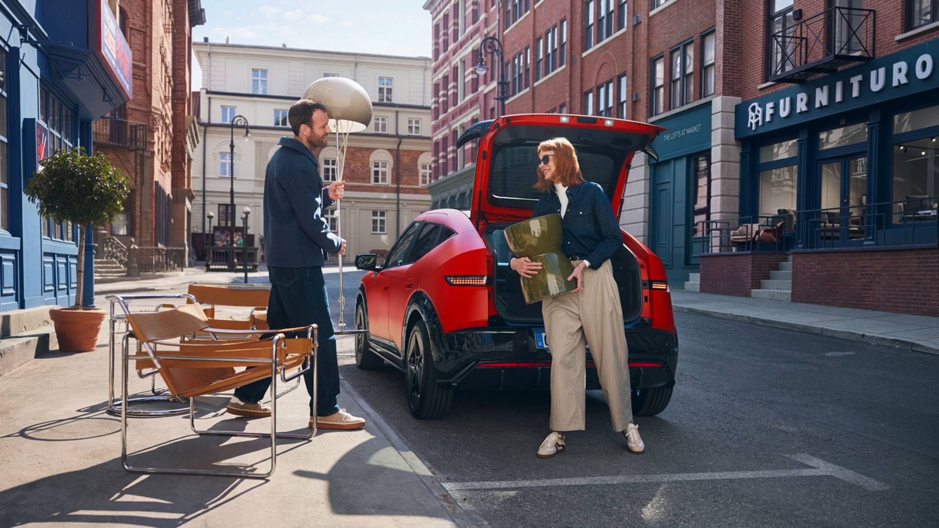 Ketten bútort pakolnak ki egy városi utcán parkoló, piros Hyundai IONIQ 3 csomagtartójából.