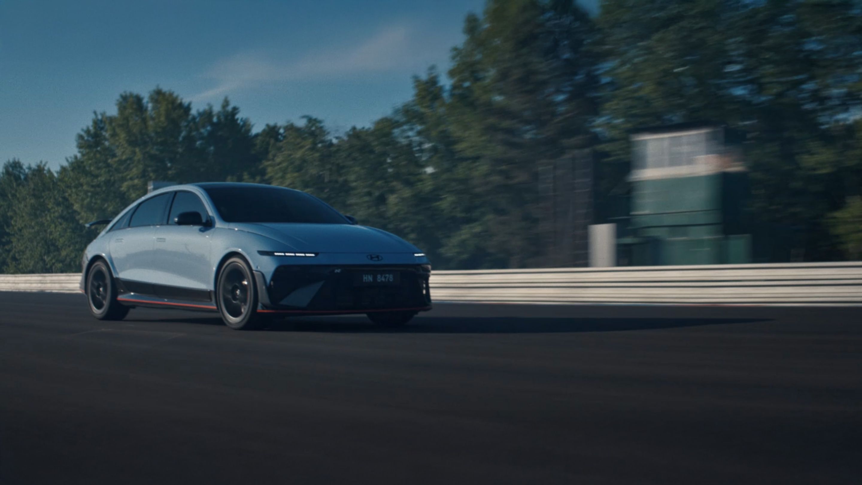 A Hyundai IONIQ 6 N elektromos sportautó versenypályán száguld, a háttérben fák és egy épület látható